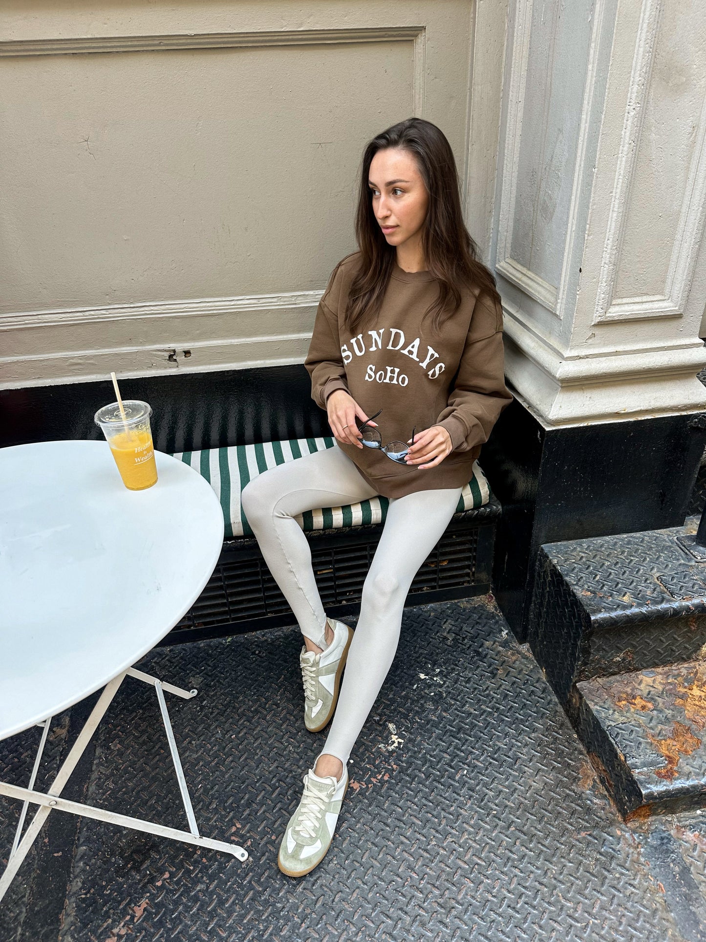 Brown crewneck sweatshirt with 'SUNDAY SoHo' logo, worn with light pants and boots.