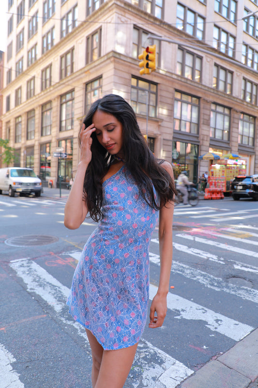 A woman wearing a blue sleeveless mini dress with a pink and white abstract pattern stands on a city crosswalk in front of a tall urban building.