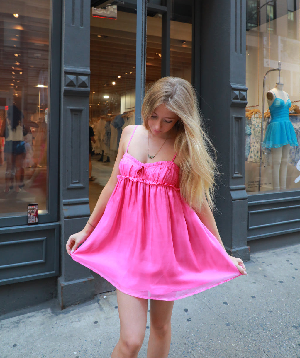 A young woman with long blonde hair stands on a city sidewalk in front of a dark storefront, wearing a bright pink sleeveless mini dress and holding it out to show its flowy shape.