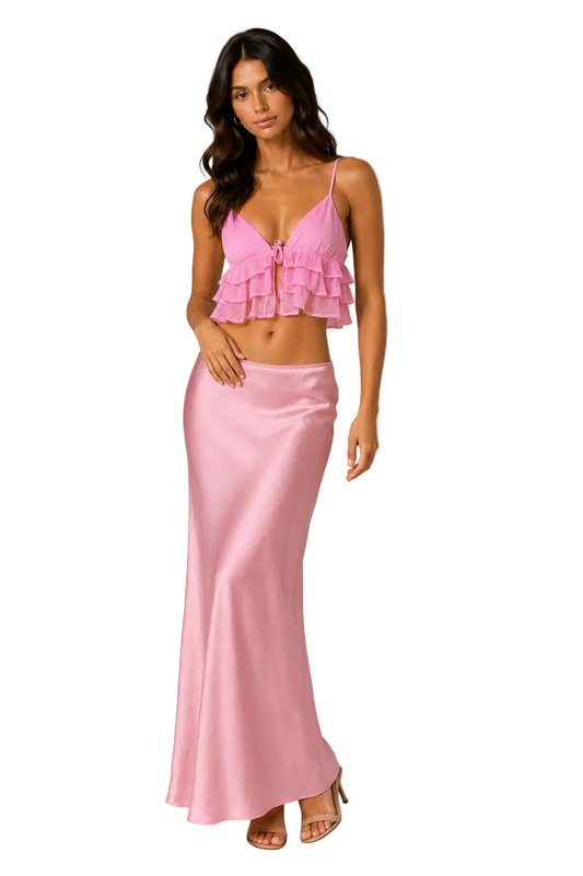 A woman modeling a two-piece pink outfit consisting of a ruffled crop top with spaghetti straps and a matching satin maxi skirt, set against a plain beige studio background.