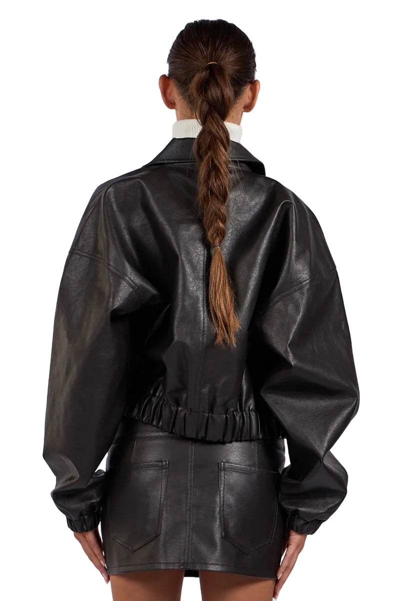 A model shown from the back wearing a black faux-leather jacket and matching mini skirt against a light gray studio background.