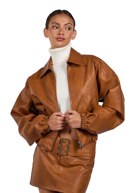 A model wearing a brown faux leather jacket and matching mini skirt over a white turtleneck sweater, posed against a light gray studio background.