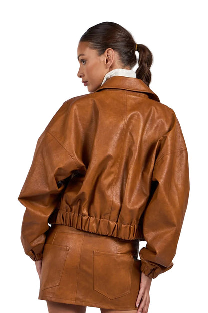 A woman shown from the back wearing a brown faux-leather jacket and matching mini skirt against a light gray studio background.