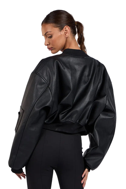 A woman shown from the back wearing a black faux-leather bomber jacket with oversized sleeves and black pants, photographed against a light gray studio background.